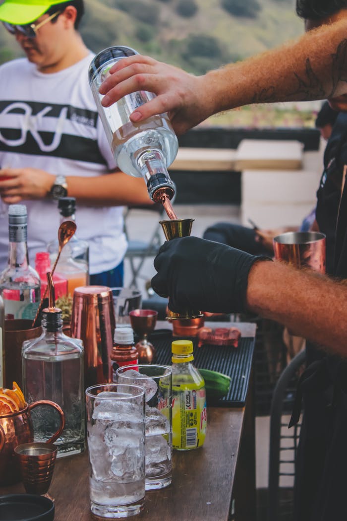 our-experience An outdoor bartender expertly pouring alcohol into a jigger amidst various bar tools and ingredients.